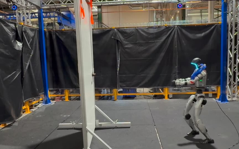 A humanoid robot approaches a door in the original Energy-Based Policy Orchestration real-robot demonstration.